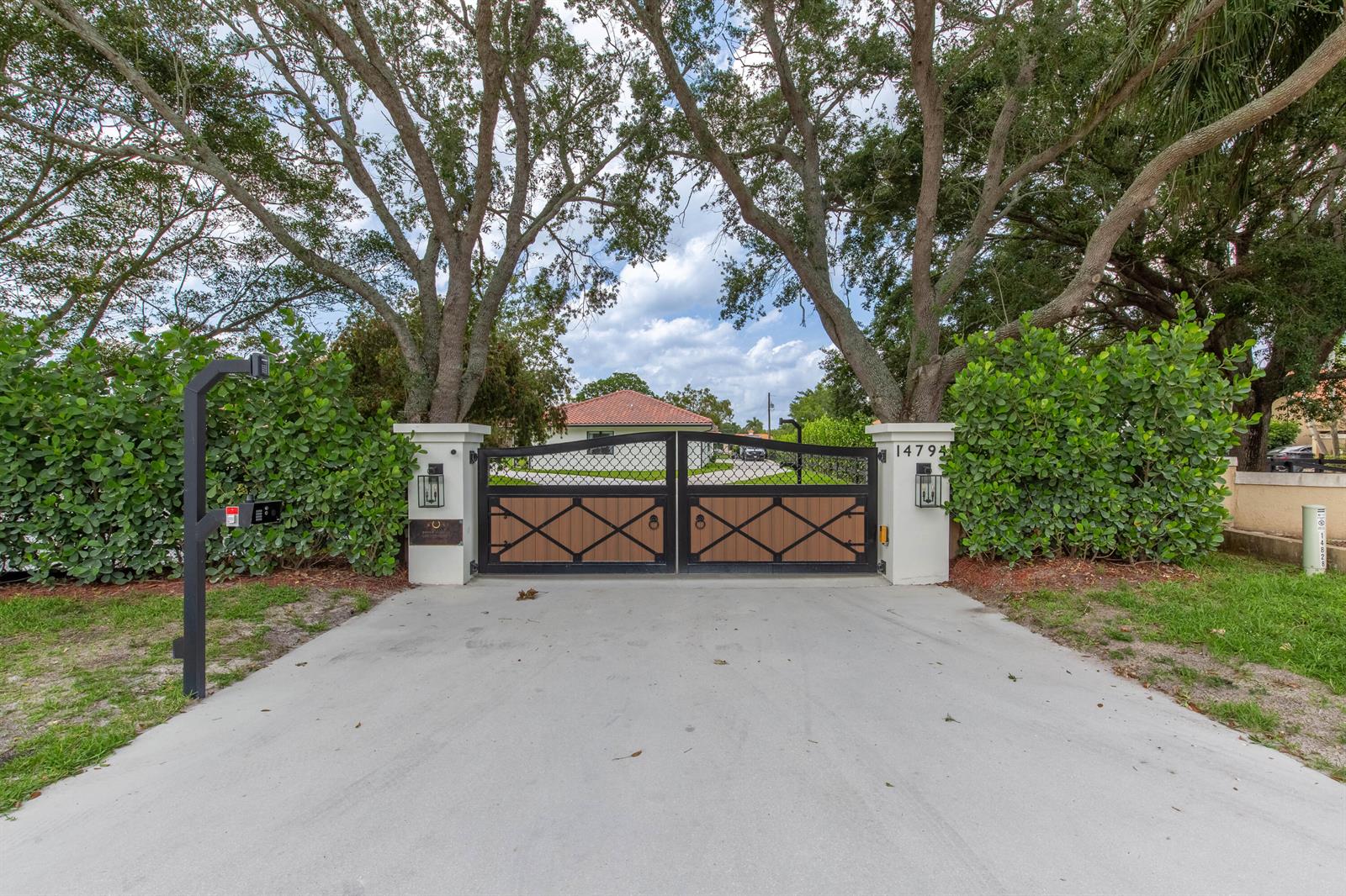 A concrete driveway leads to a gated entrance with white pillars and mature trees — 14794 Rolling Rock Place, Wellington FL 33414, WELLINGTON, FL