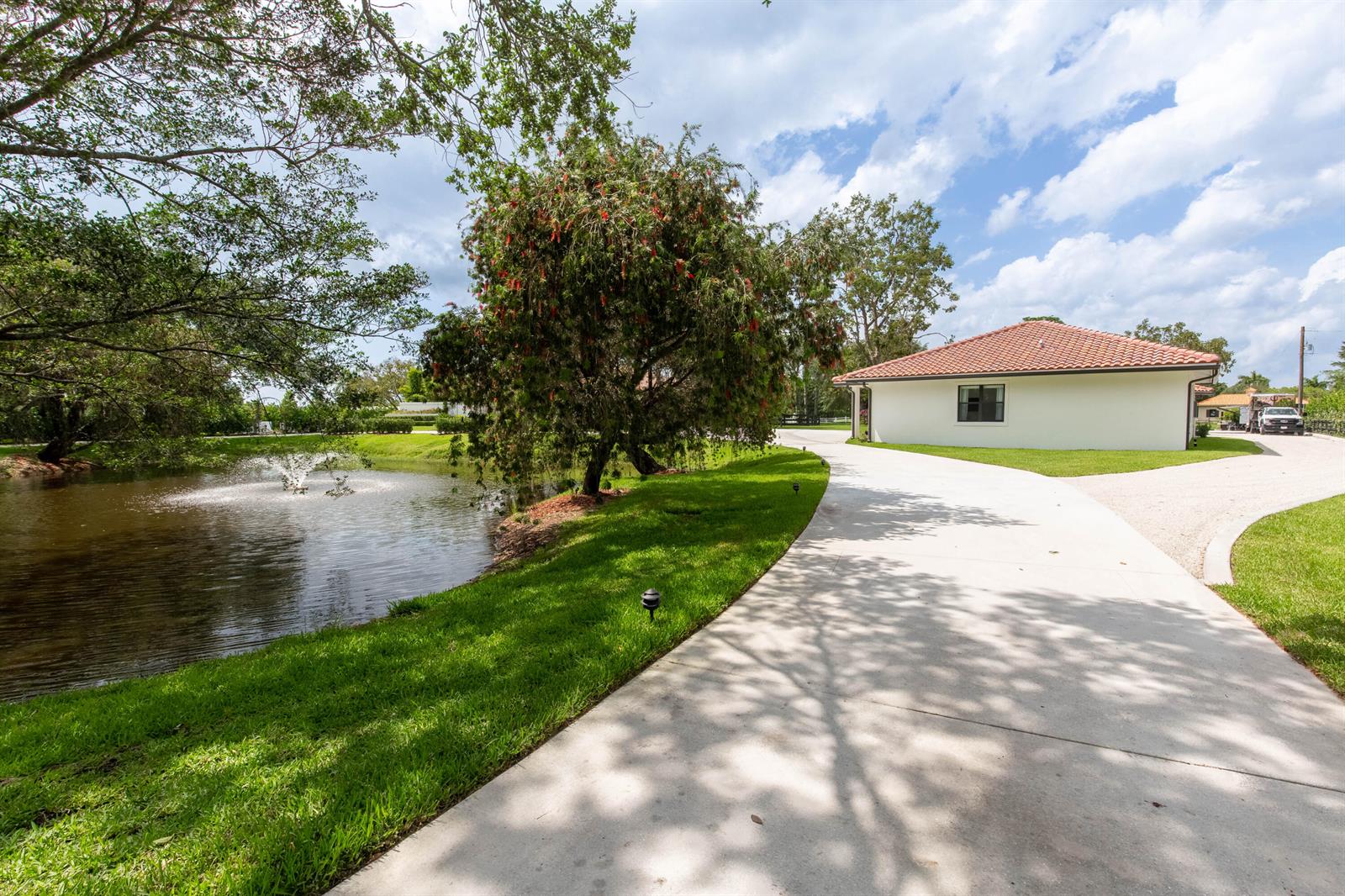 A concrete driveway leads past a pond with a fountain toward a white house — 14794 Rolling Rock Place, Wellington FL 33414, WELLINGTON, FL