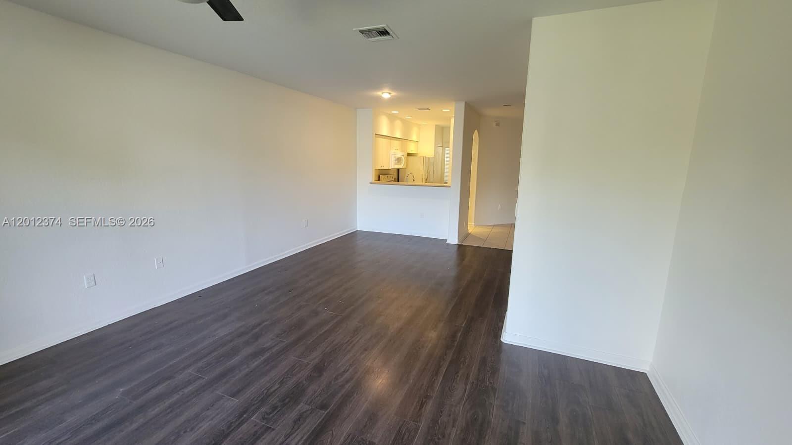 An empty living room features dark wood-look flooring and an open pass-through to the kitchen — 8353 SW 26th St # 104, Miramar FL 33025, MIRAMAR, FL
