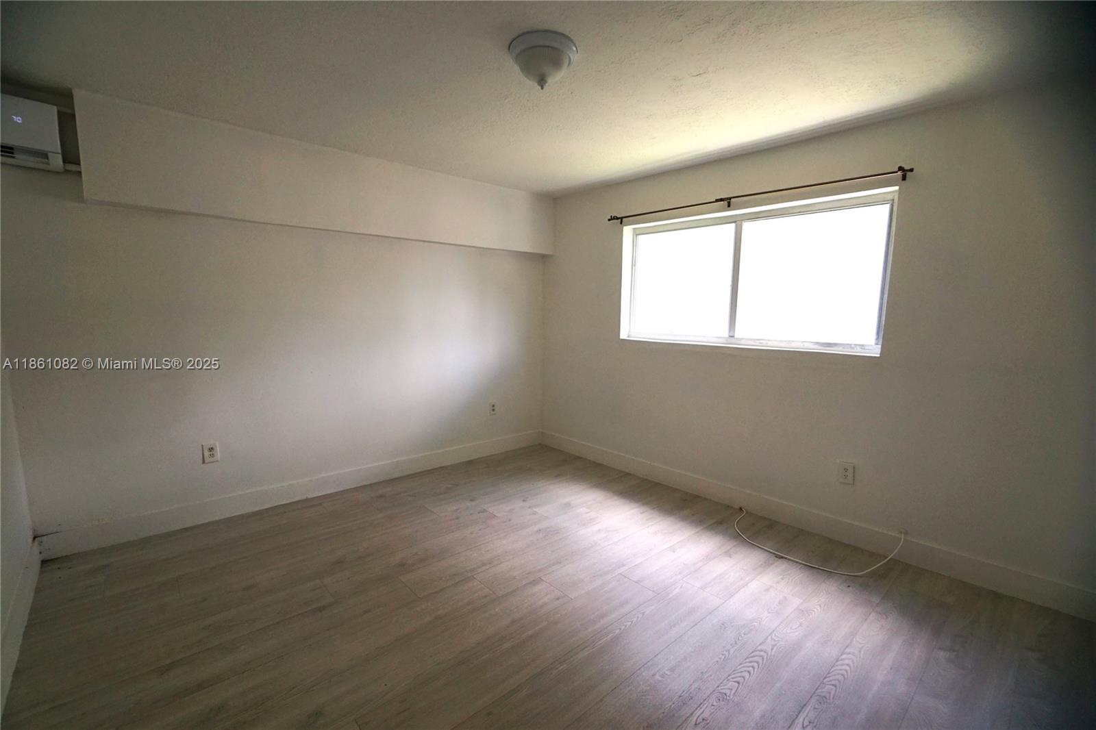 An empty bedroom features light wood-look flooring, white walls, a window, and a wall-mounted air conditioner — 584 NW 65th St # 3, Miami FL 33150, MIAMI, FL