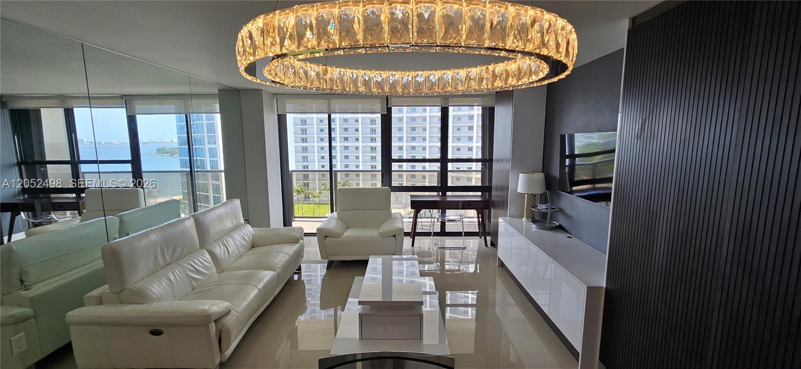 A modern living room features a white leather sofa, a crystal chandelier, and floor-to-ceiling windows — 600 NE 36th St # 1509, Miami FL 33137, MIAMI, FL