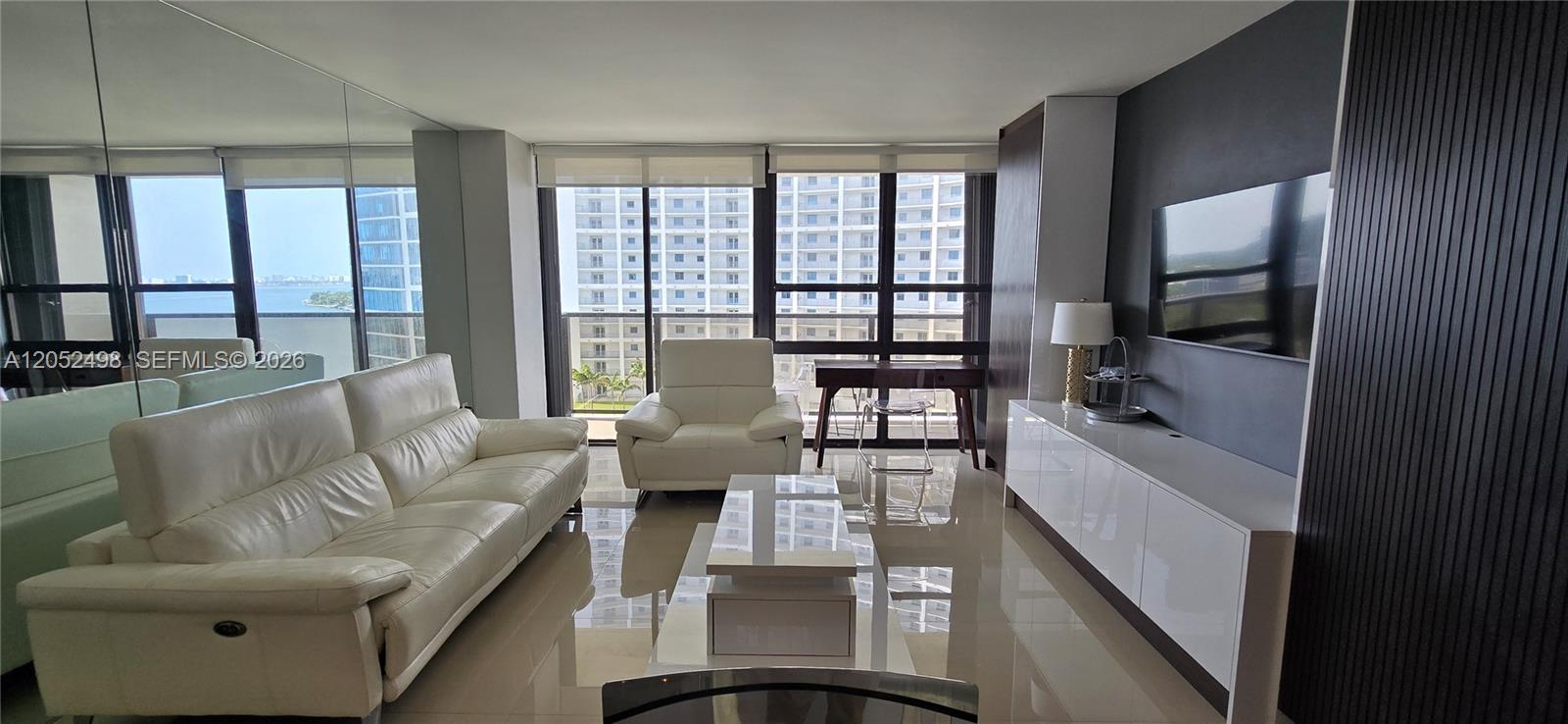 A modern living room features white leather furniture, a glossy console, and floor-to-ceiling windows — 600 NE 36th St # 1509, Miami FL 33137, MIAMI, FL