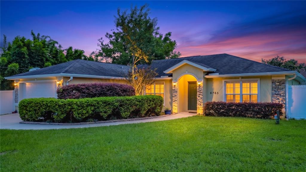 A single-story house with a tan exterior, stone accents, and a manicured hedge at dusk — 6765 NW 14TH AVENUE, OCALA, FL