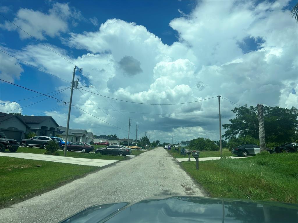 A paved street runs through a residential neighborhood under a large, cloudy blue sky — 1219 NW 24TH PLACE, CAPE CORAL, FL