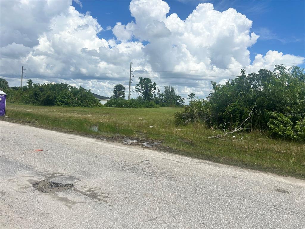 A vacant, grassy lot sits along an asphalt road under a cloudy blue sky — 1219 NW 24TH PLACE, CAPE CORAL, FL