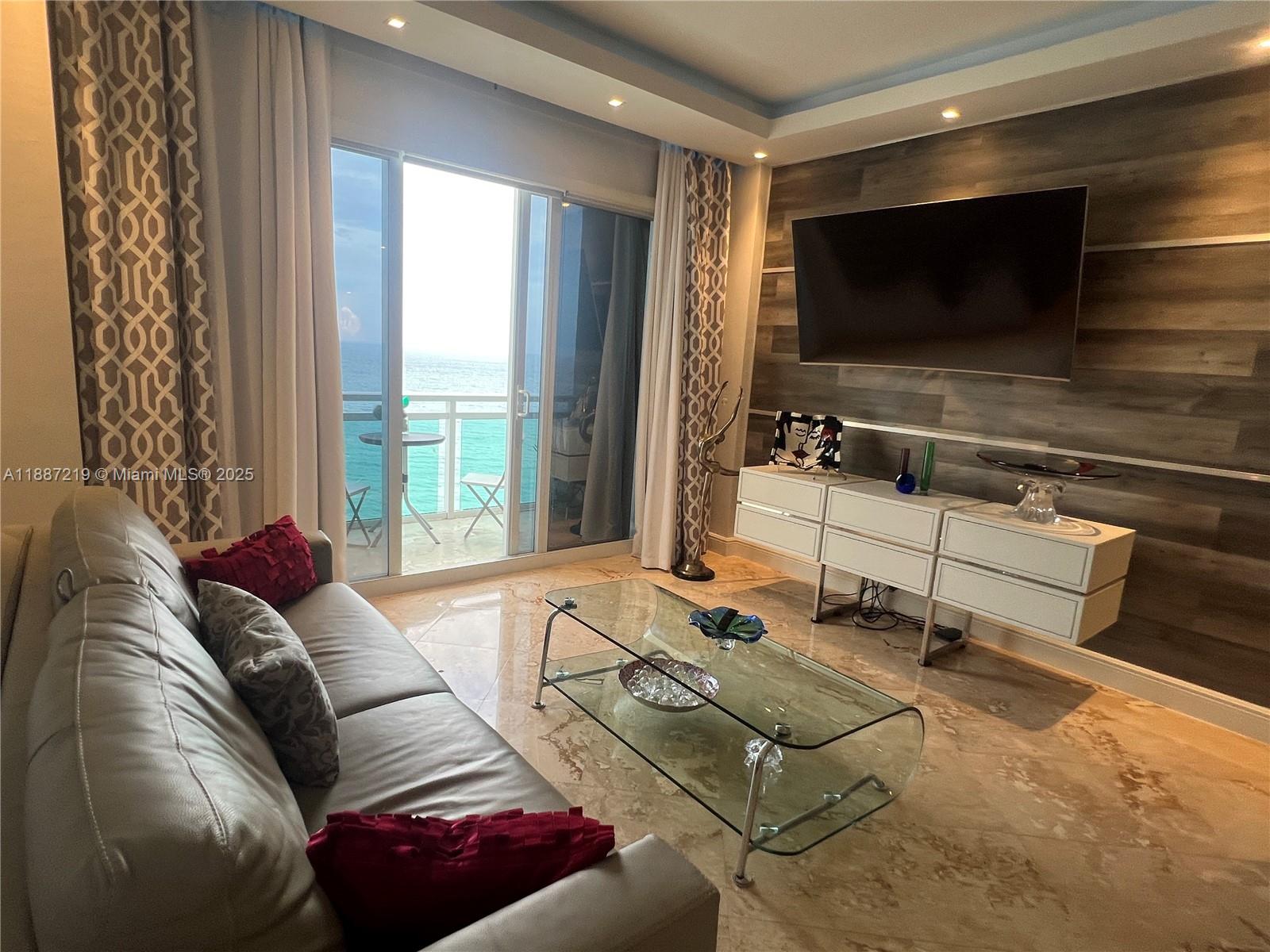 A living room features a glass coffee table, a wall-mounted television, and a balcony view — 6917 Collins Ave, Miami Beach FL 33141, MIAMI BEACH, FL