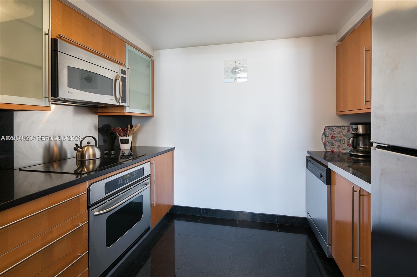 A galley kitchen features light wood cabinets, black countertops, and stainless steel appliances — 6365 Collins Ave # 1811, Miami Beach FL 33141, MIAMI BEACH, FL