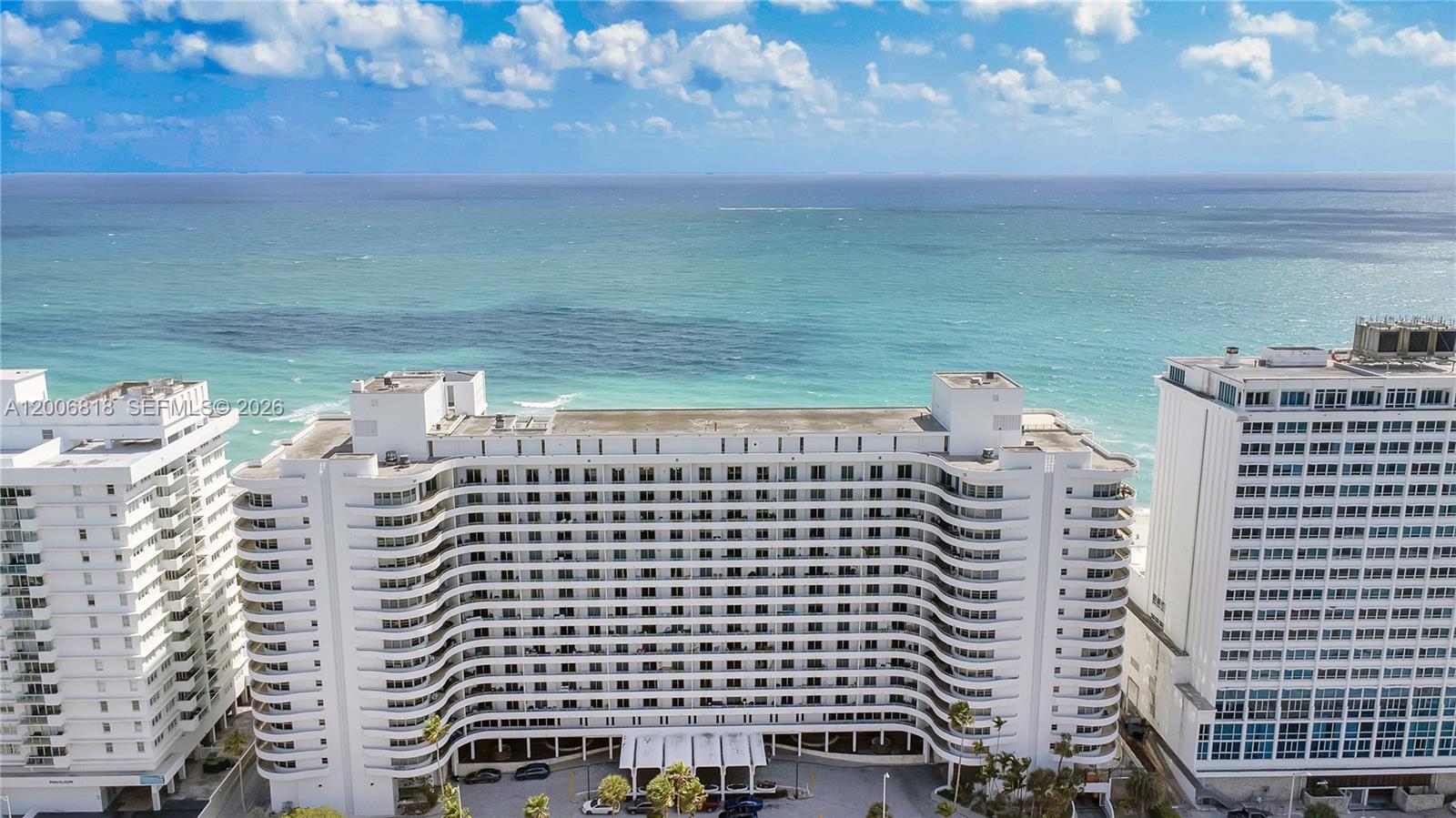An aerial view shows a white, curved beachfront apartment building with multiple balconies — 5555 Collins Ave # 16A, Miami Beach FL 33140, MIAMI BEACH, FL