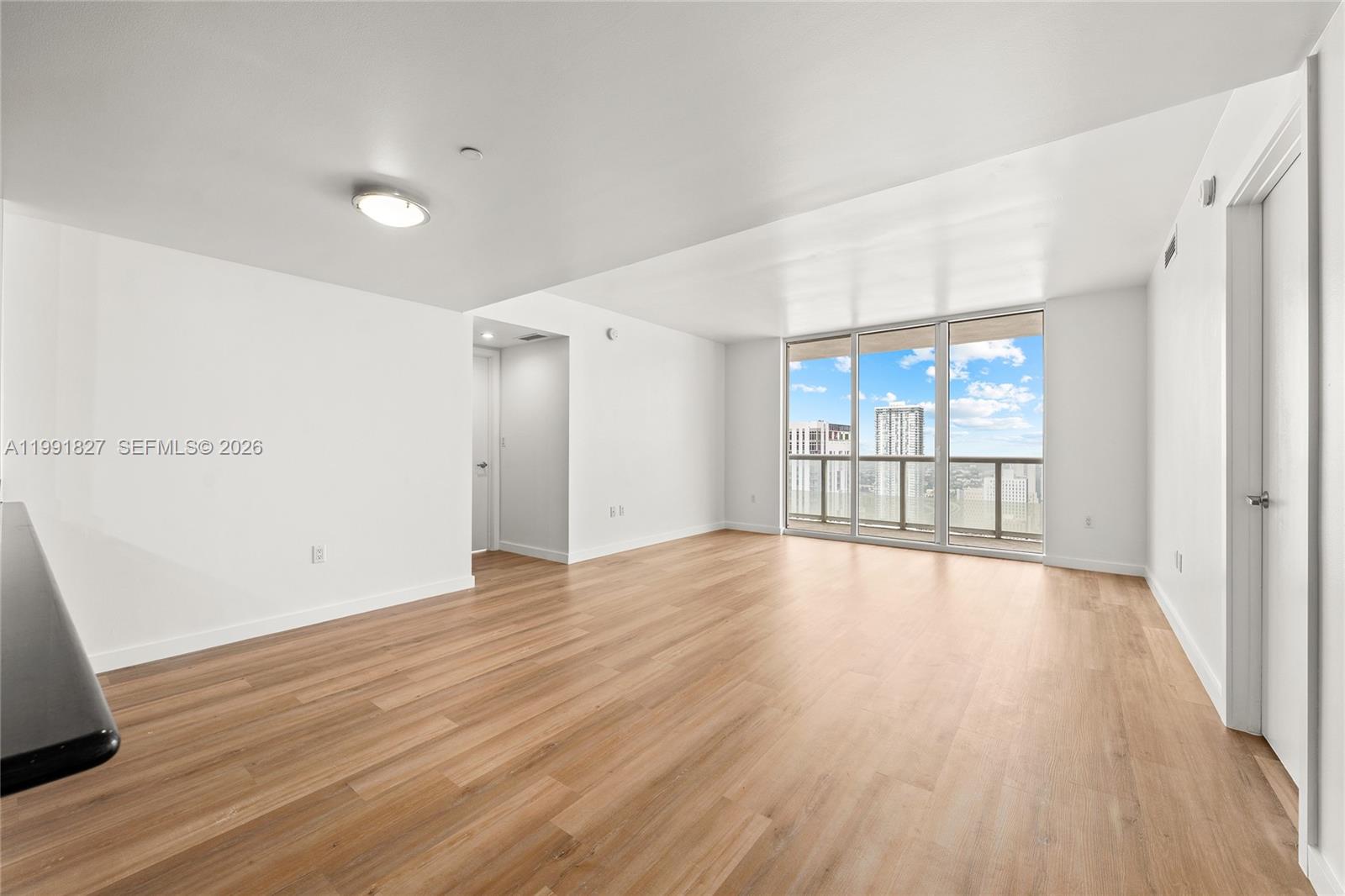An empty living room features light wood-look flooring, white walls, and a balcony with city views — 50 Biscayne Blvd # 4111, Miami FL 33132, MIAMI, FL