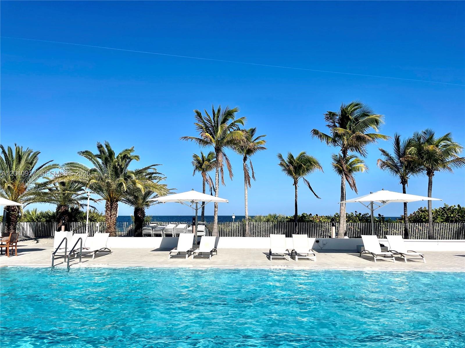 A swimming pool with lounge chairs and umbrellas sits before palm trees and the ocean — 607 Ocean Dr # 3K, Key Biscayne FL 33149, KEY BISCAYNE, FL
