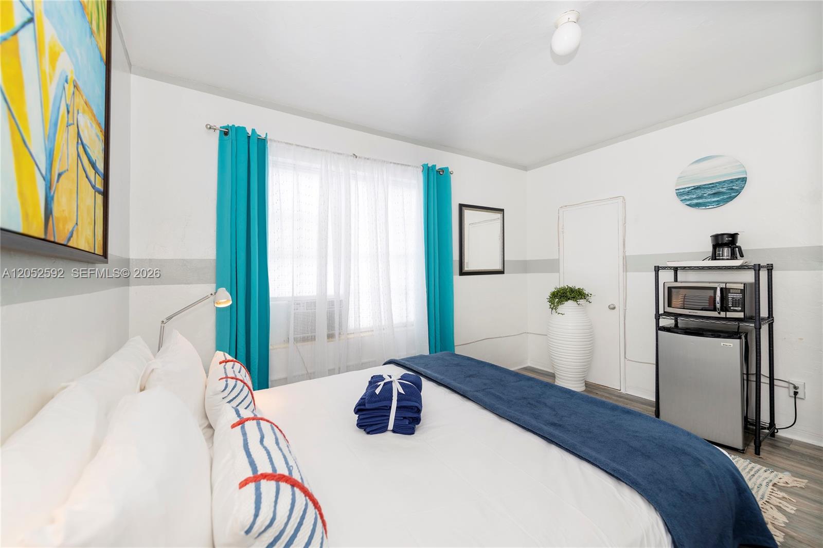 A bedroom features a bed with blue accents, turquoise curtains, and a small kitchenette area — 6760 Collins Ave # 29, Miami Beach FL 33141, MIAMI BEACH, FL