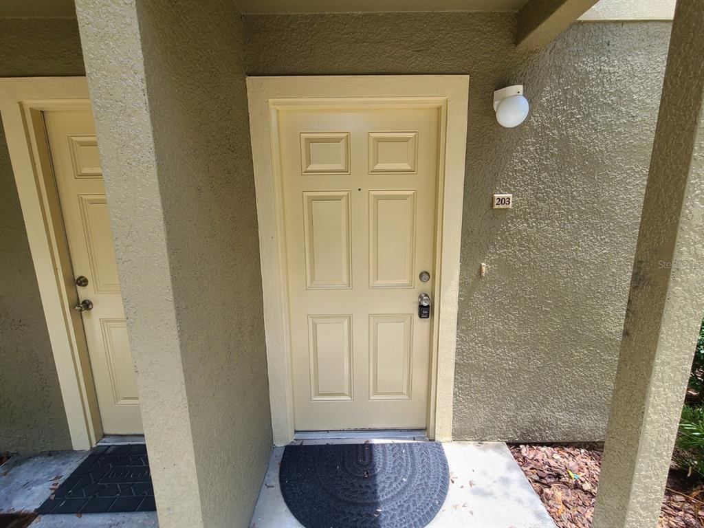 A beige exterior door with a lockbox and a welcome mat at the entrance — 36122 LAKE CHASE BOULEVARD UNIT 203, ZEPHYRHILLS, FL