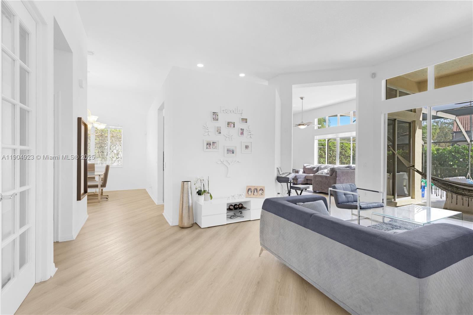 A bright living room features light wood flooring, a grey sofa, and a family photo wall — 1102 Camellia Cir, Weston FL 33326, WESTON, FL