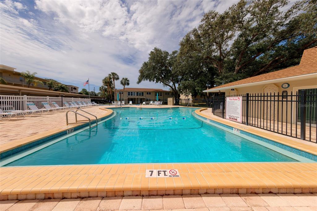 An outdoor swimming pool with a tiled deck and surrounding apartment buildings under a cloudy sky — 946 VIRGINIA STREET UNIT 107, DUNEDIN, FL