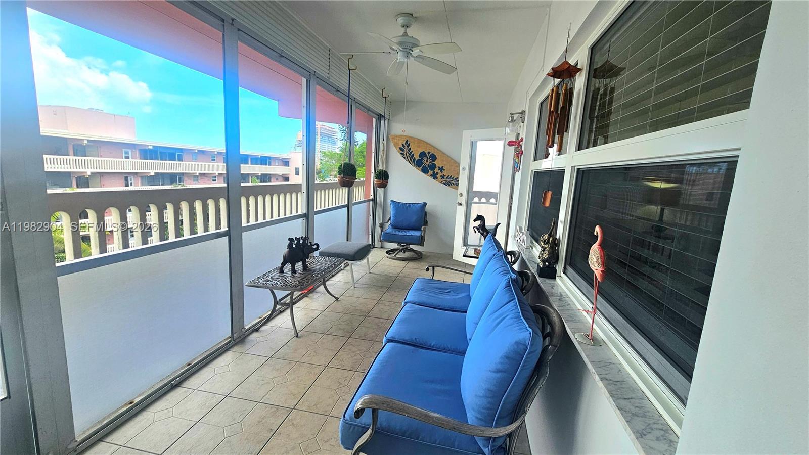 A screened-in balcony features blue patio furniture, a surfboard wall decoration, and tiled flooring — 180 NE 12th Ave # 10E, Hallandale Beach FL 33009, HALLANDALE BEACH, FL
