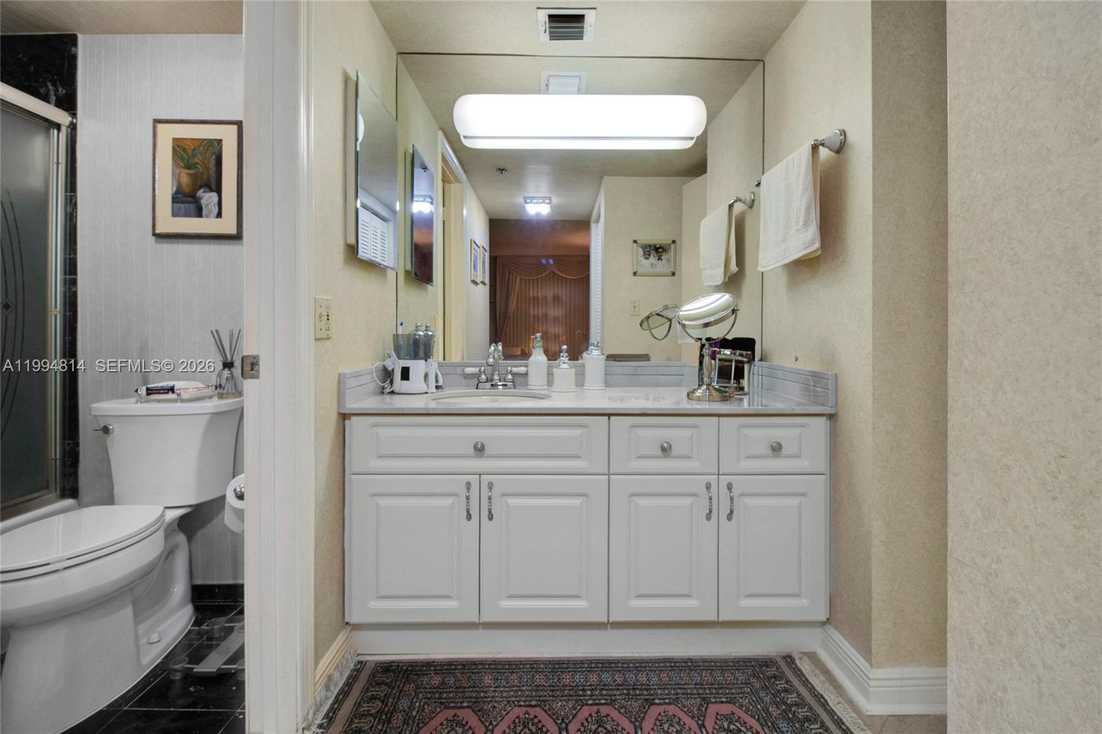 A bathroom features a white vanity with a marble countertop, a large mirror, and toilet — 6767 Collins Ave # 504, Miami Beach FL 33141, MIAMI BEACH, FL
