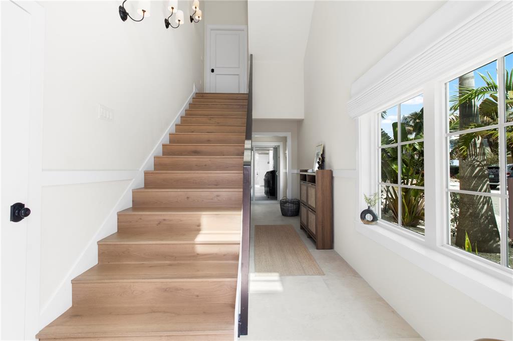 A bright entryway features a wooden staircase, white walls, and a window overlooking palm trees — 416 ALAMANDA ROAD, ANNA MARIA, FL