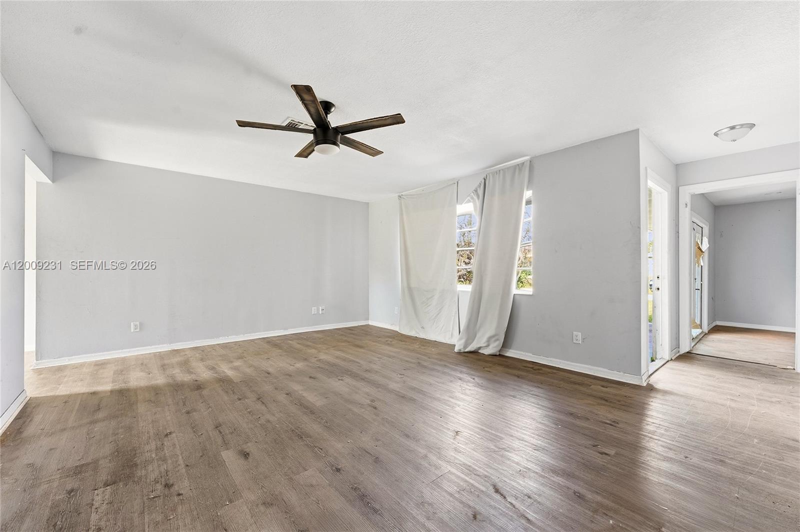 An empty room features light gray walls, wood-look flooring, a ceiling fan, and white curtains — 5808 Spruce Dr, Fort Pierce FL 34982, FORT PIERCE, FL