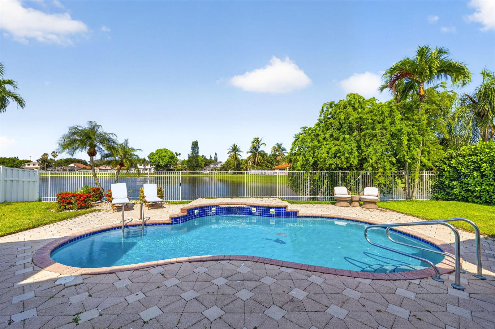 A kidney-shaped swimming pool with a brick paver deck overlooks a lake and white fence — 912 NW 132nd Avenue, Sunrise FL 33325, SUNRISE, FL