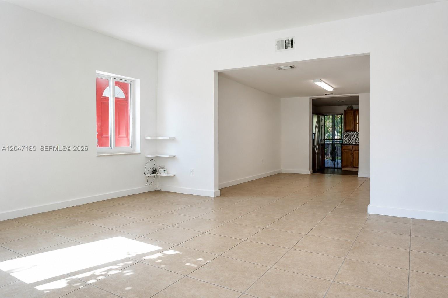 An empty room with white walls, beige tile flooring, and a view into a kitchen — 861 W 32nd St, Hialeah FL 33012, HIALEAH, FL