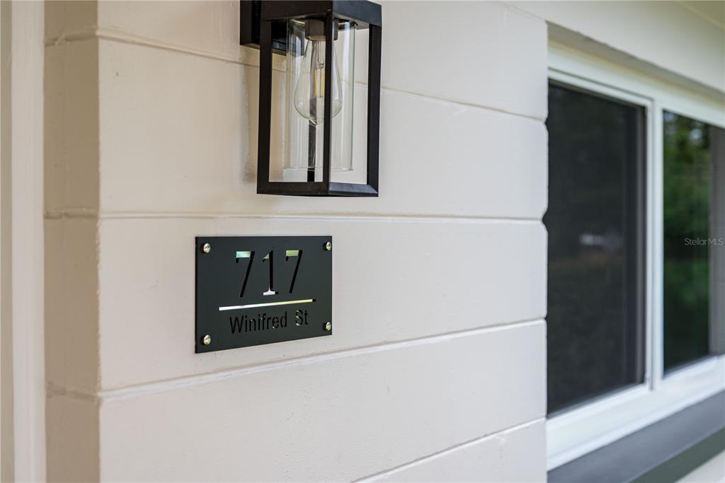 A modern house number plaque is mounted on a white horizontal-sided exterior wall — 717 W WINIFRED STREET, AVON PARK, FL