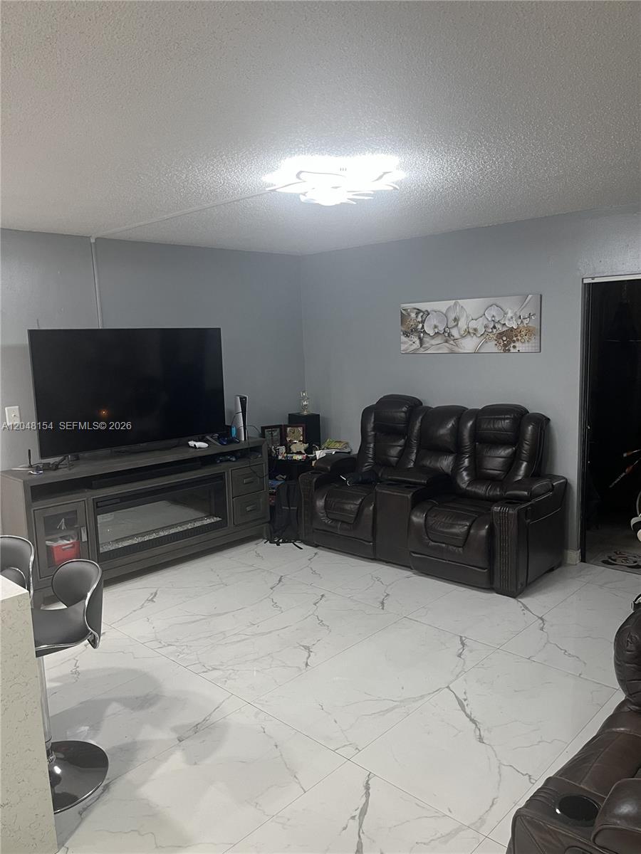 A living room features a dark leather sofa, a television console with a fireplace, and white marble-patterned tile flooring — 819 NE 199th St # 101, Miami FL 33179, MIAMI, FL