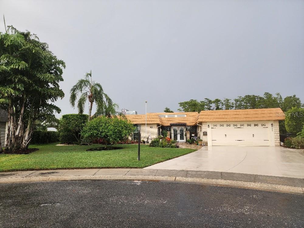 A single-story house with a tan tile roof and a two-car garage sits behind a lawn — 1411 JACOBSON CIRCLE, SUN CITY CENTER, FL