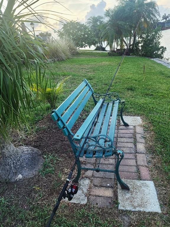 A teal metal bench sits on a brick path near a grassy waterfront area — 1411 JACOBSON CIRCLE, SUN CITY CENTER, FL