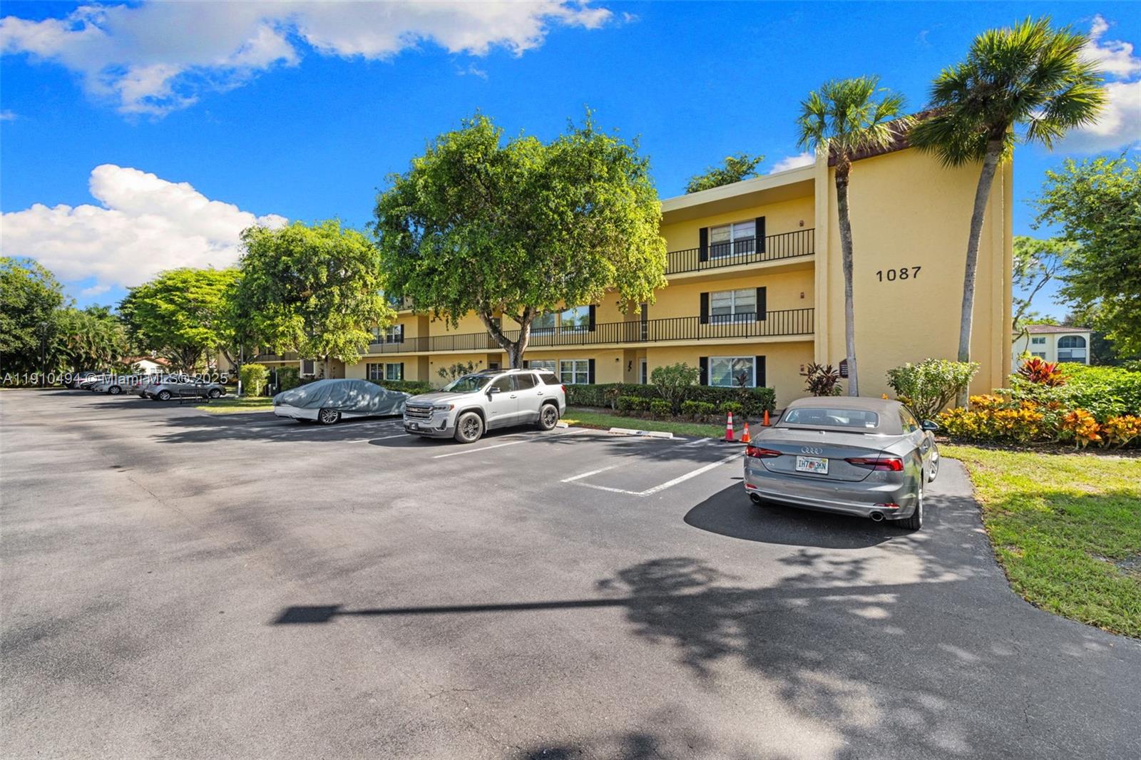 A three-story yellow apartment building stands behind a parking lot with several parked cars — 1087 Forest Lakes Drive # 309, Naples FL 34105, NAPLES, FL