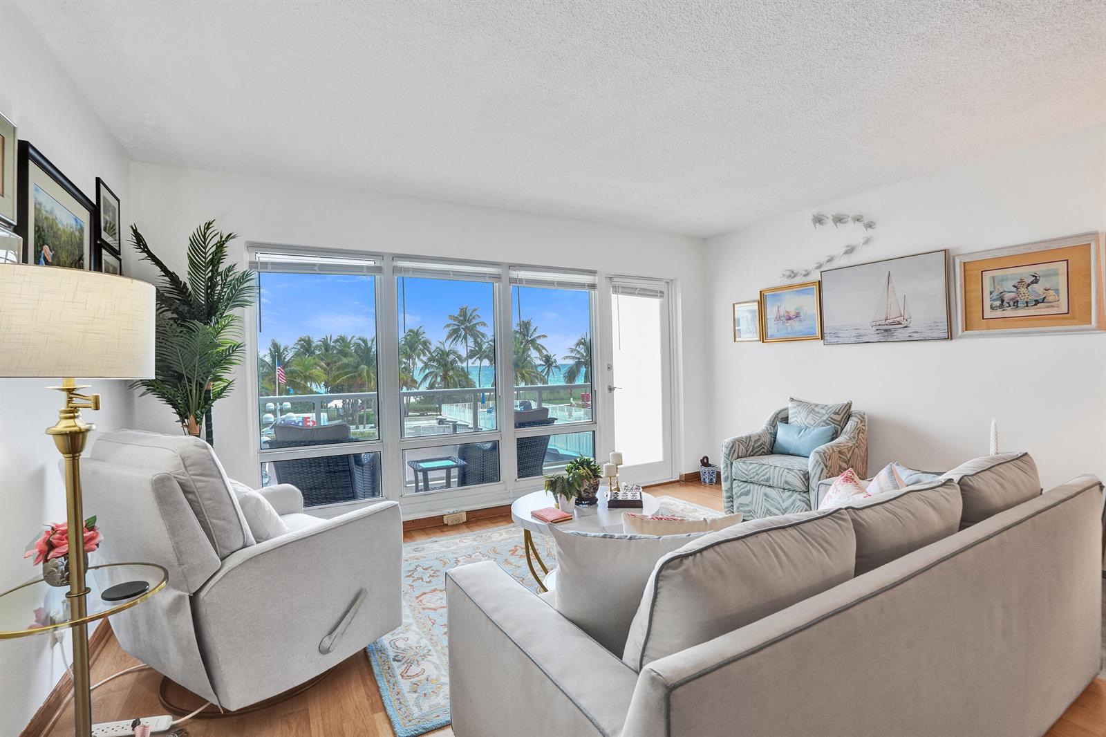 A living room features light-colored furniture, a round coffee table, and large windows overlooking palm trees — 111 N Pompano Beach Boulevard # 411, Pompano Beach FL 33062, POMPANO BEACH, FL