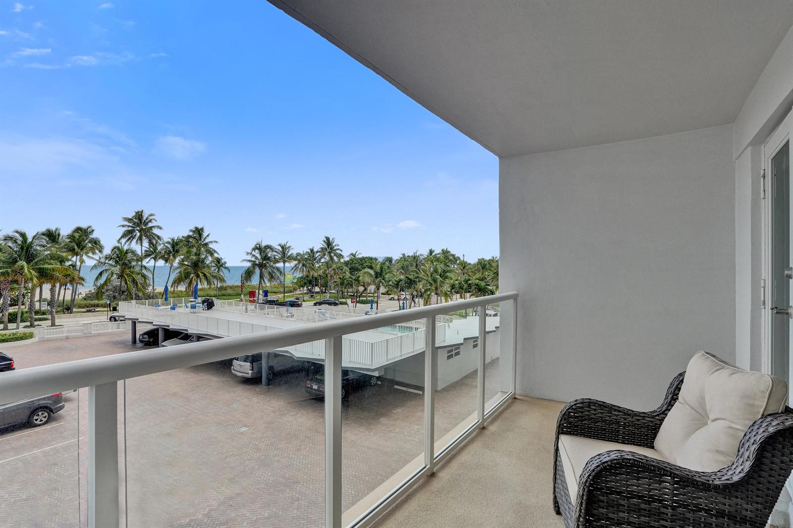 A balcony with a wicker chair overlooks a parking lot, palm trees, and the ocean — 111 N Pompano Beach Boulevard # 411, Pompano Beach FL 33062, POMPANO BEACH, FL
