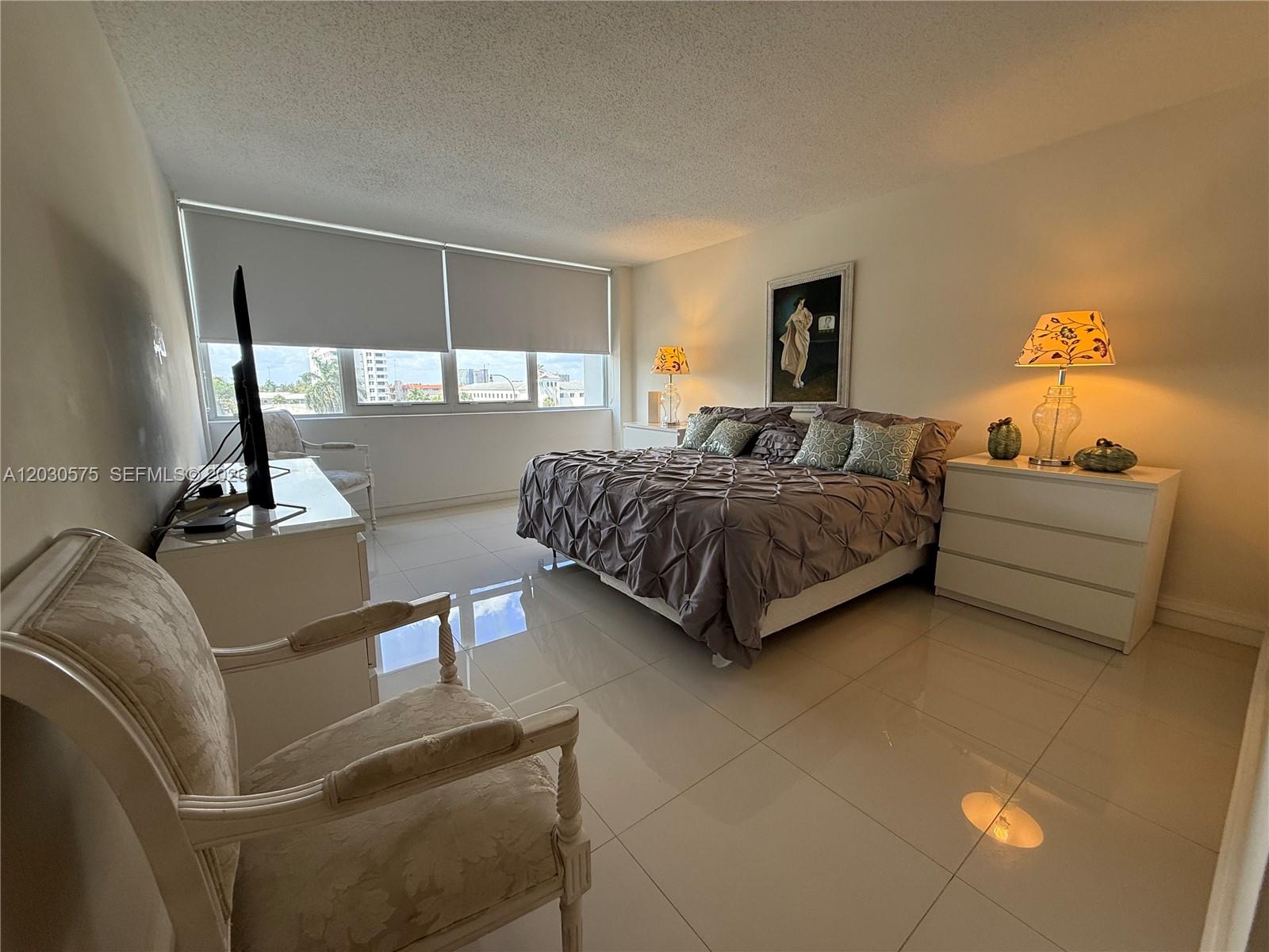 A bedroom features a bed with a gray comforter, white tile floors, and large windows — 3140 S Ocean Dr # 310, Hallandale Beach FL 33009, HALLANDALE BEACH, FL