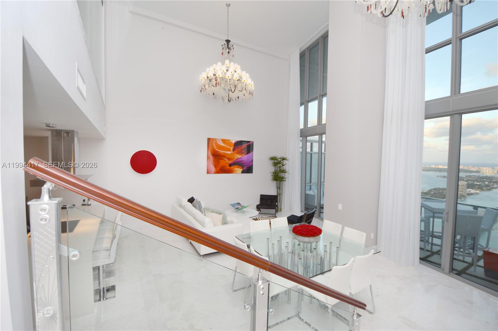 A high-ceilinged living and dining area features a glass table, chandelier, and floor-to-ceiling windows — 1100 Biscayne Blvd # 6104, Miami FL 33132, MIAMI, FL