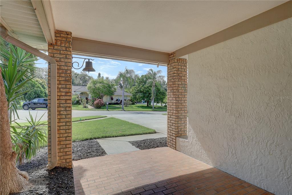 A covered brick porch looks out onto a suburban street with a hanging bell feature — 7600 DREW OAK DRIVE, SEMINOLE, FL