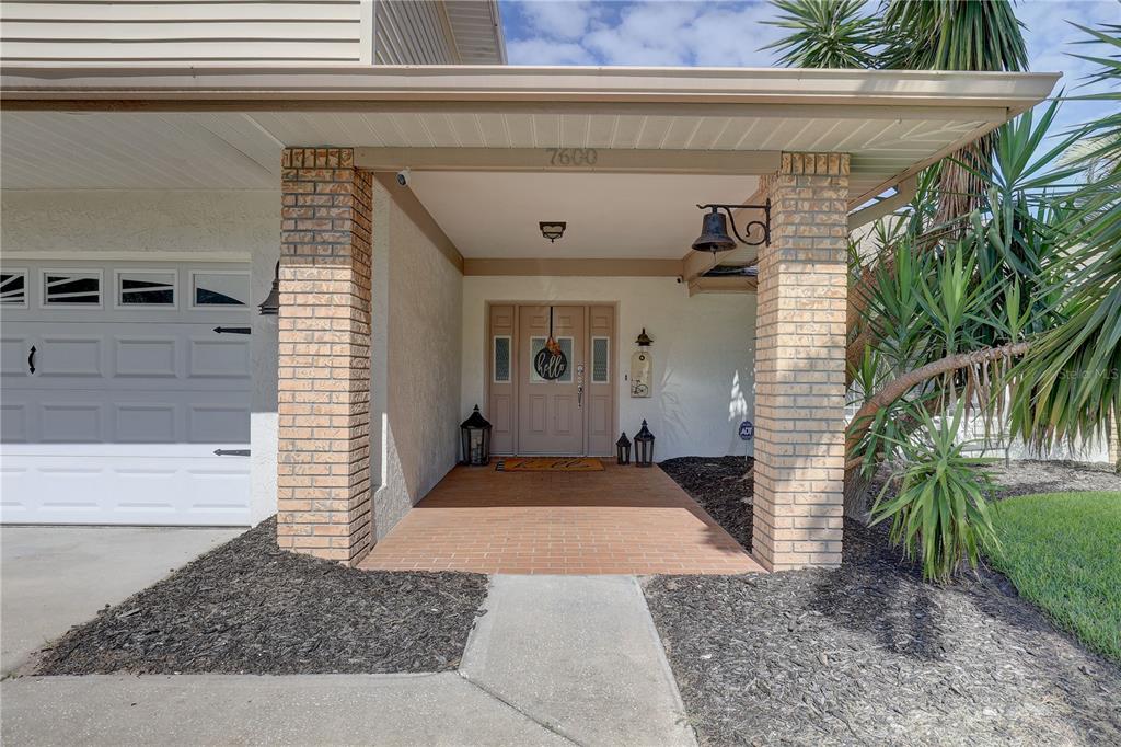 A covered brick entryway leads to a tan front door with a decorative welcome sign — 7600 DREW OAK DRIVE, SEMINOLE, FL