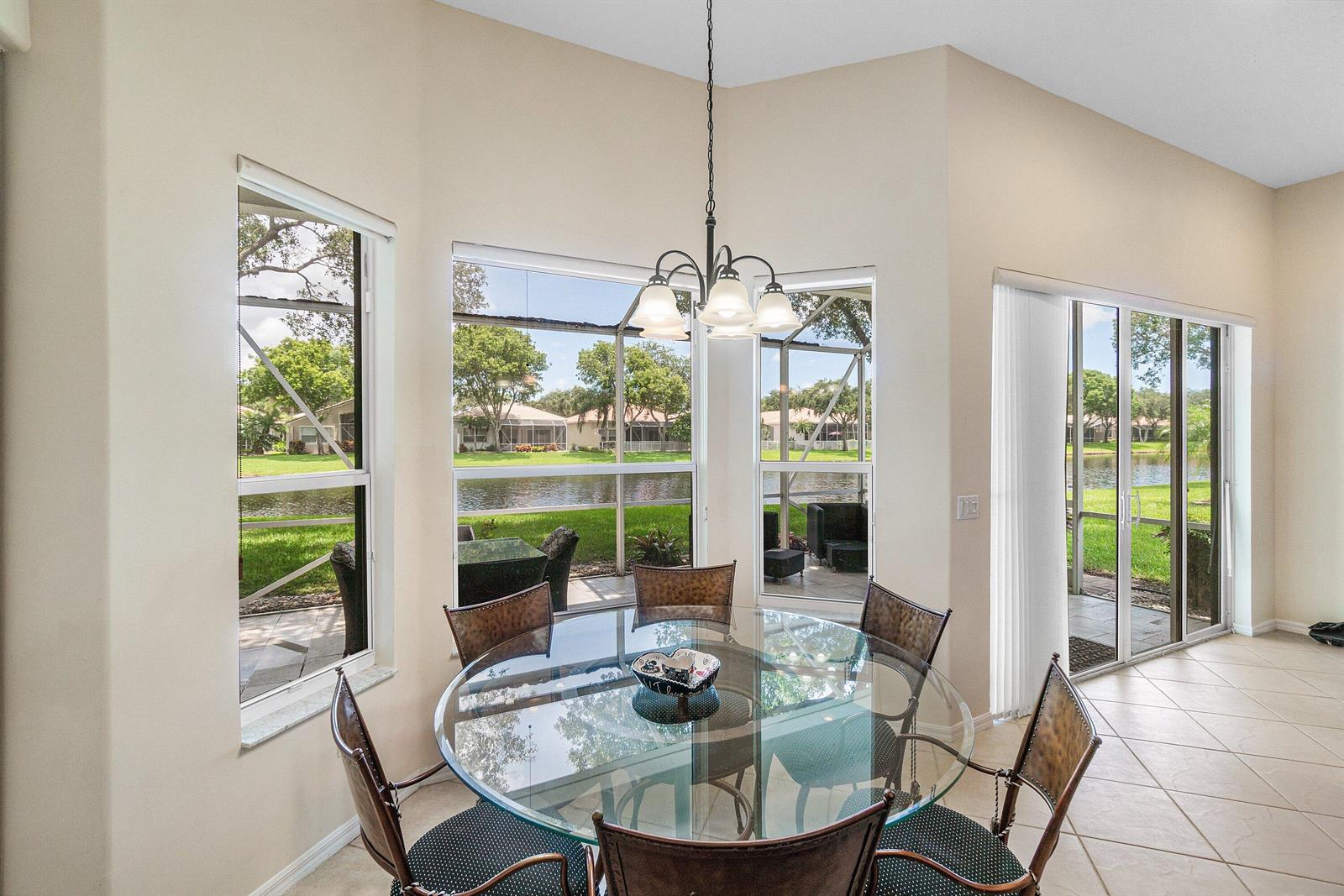 A dining area features a glass table, metal chairs, a chandelier, and large windows overlooking water — 12196 Landrum Way, Boynton Beach FL 33437, BOYNTON BEACH, FL