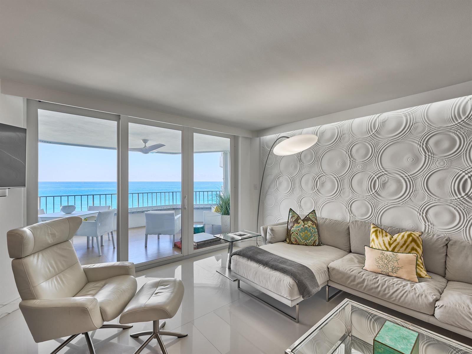 A modern living room features a textured accent wall, white tile floors, and ocean views — 550 S Ocean Boulevard # 1405, Boca Raton FL 33432, BOCA RATON, FL