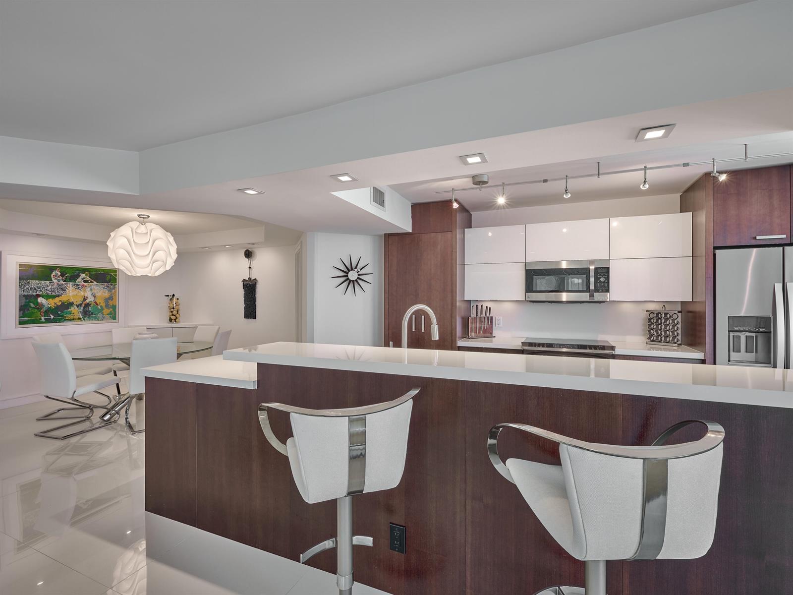 A modern kitchen features dark wood cabinetry, white countertops, and two white bar stools — 550 S Ocean Boulevard # 1405, Boca Raton FL 33432, BOCA RATON, FL