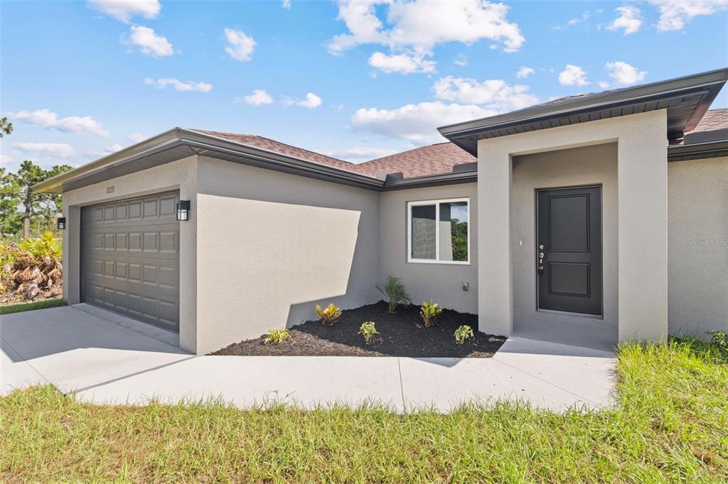A single-story house exterior features a gray stucco facade, a dark garage door, and landscaping — 12233 WAGNER LANE, PORT CHARLOTTE, FL