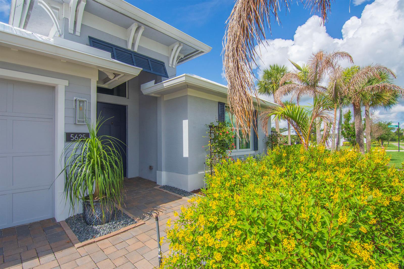 A gray house exterior features a paved walkway, a front door, and yellow flowering bushes — 5625 Arabella Lane, Vero Beach FL 32967, VERO BEACH, FL