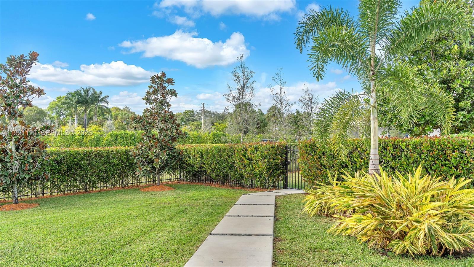 A paved walkway leads through a grassy backyard toward a gate in a tall hedge — 16772 Botaniko Dr S, Weston FL 33326, WESTON, FL