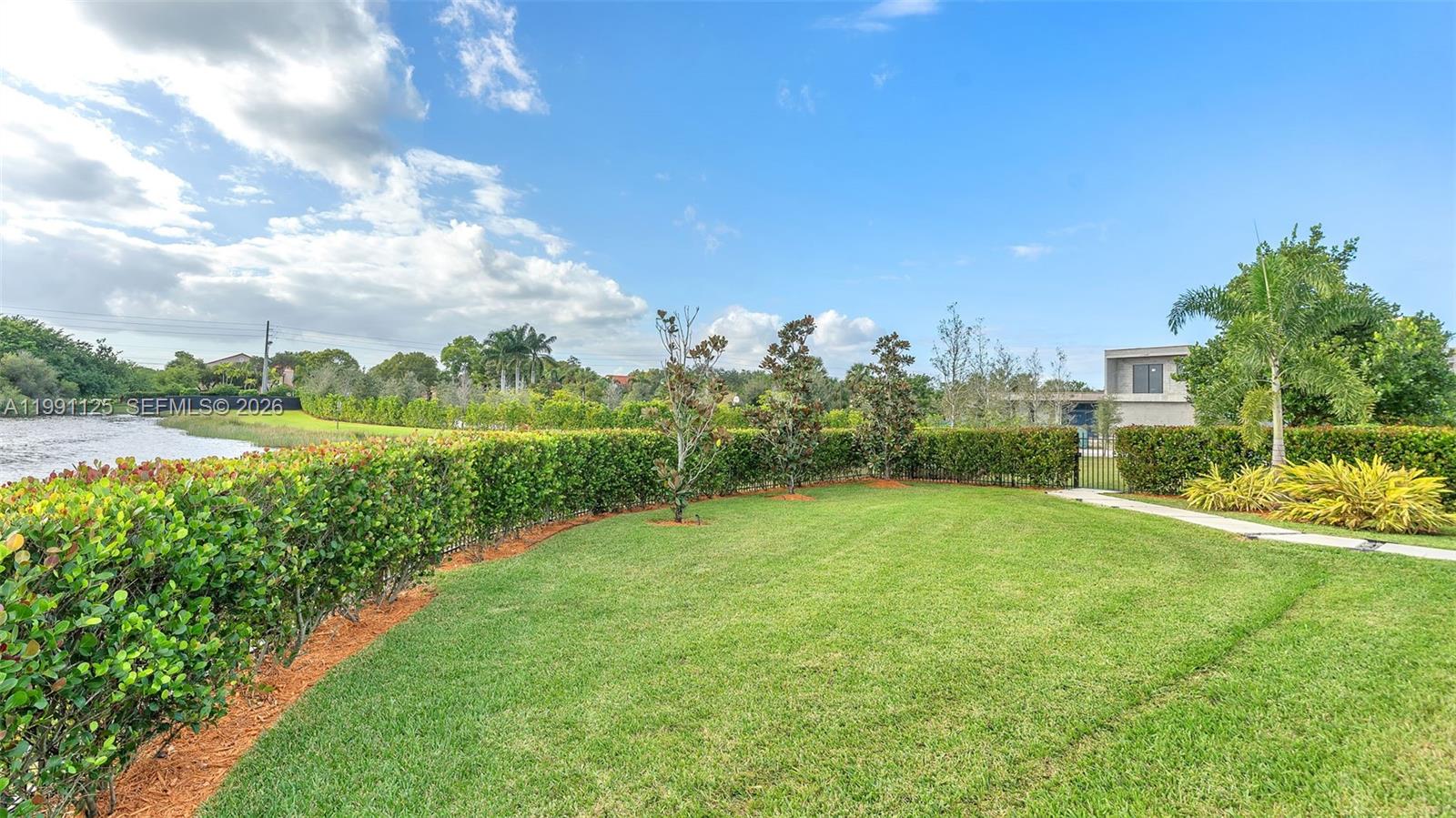 A grassy backyard features a manicured hedge, a stone path, and a view of water — 16772 Botaniko Dr S, Weston FL 33326, WESTON, FL