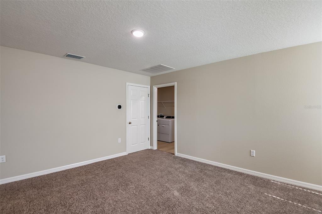 A carpeted room with beige walls, a white door, and an open doorway to a laundry area — 9110 BELLEVIEW FOREST PASS, DADE CITY, FL
