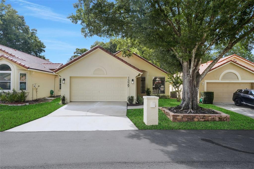 A single-story house with a beige stucco exterior, a two-car garage, and a large tree — 1329 AUGUSTA NATIONAL BOULEVARD, WINTER SPRINGS, FL