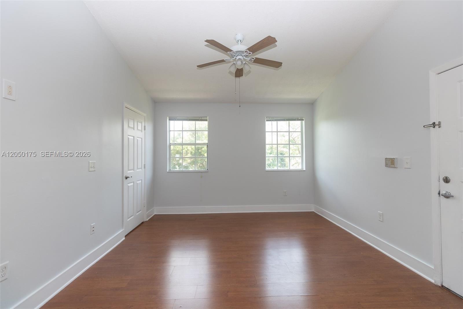 An empty room features light gray walls, wood-look flooring, two windows, and a ceiling fan — 18308 NW 68th Ave # L, Hialeah FL 33015, HIALEAH, FL