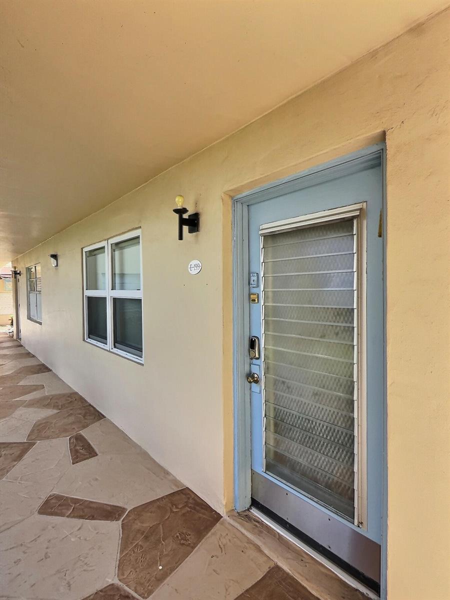 A light blue door with a louvered window sits on a walkway next to a window — 199 Burgundy E, Delray Beach FL 33484, DELRAY BEACH, FL