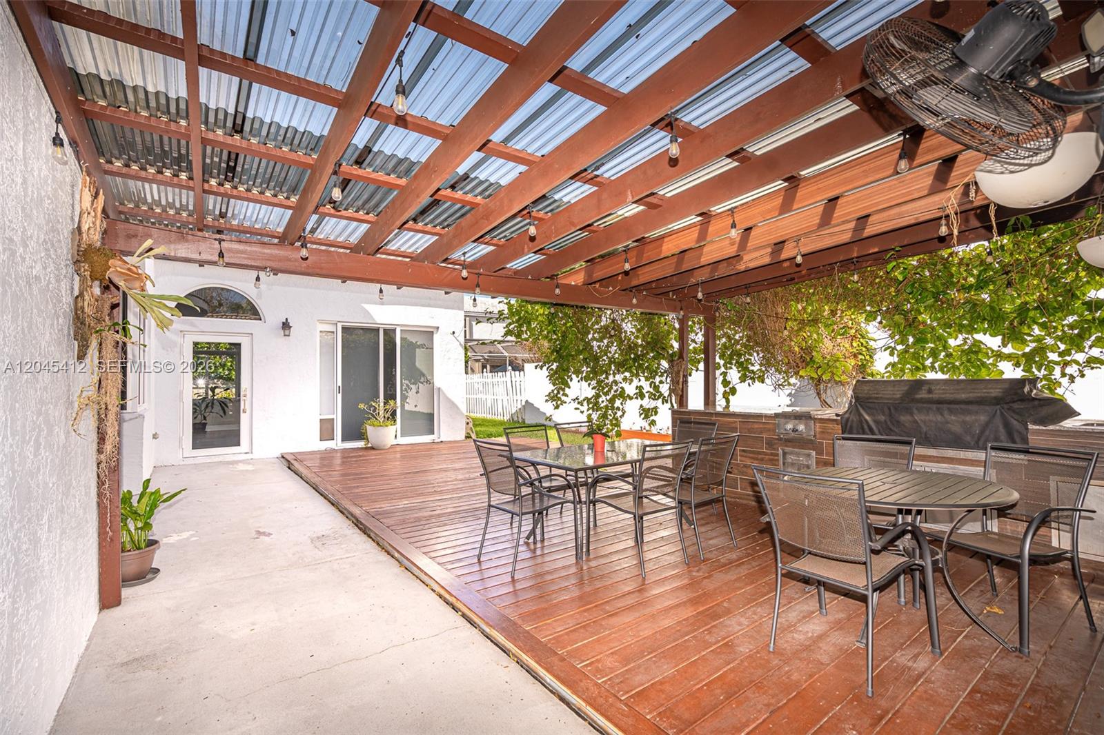 A covered wooden deck features outdoor dining sets, a built-in grill, and string lighting — PEMBROKE PINES, FL