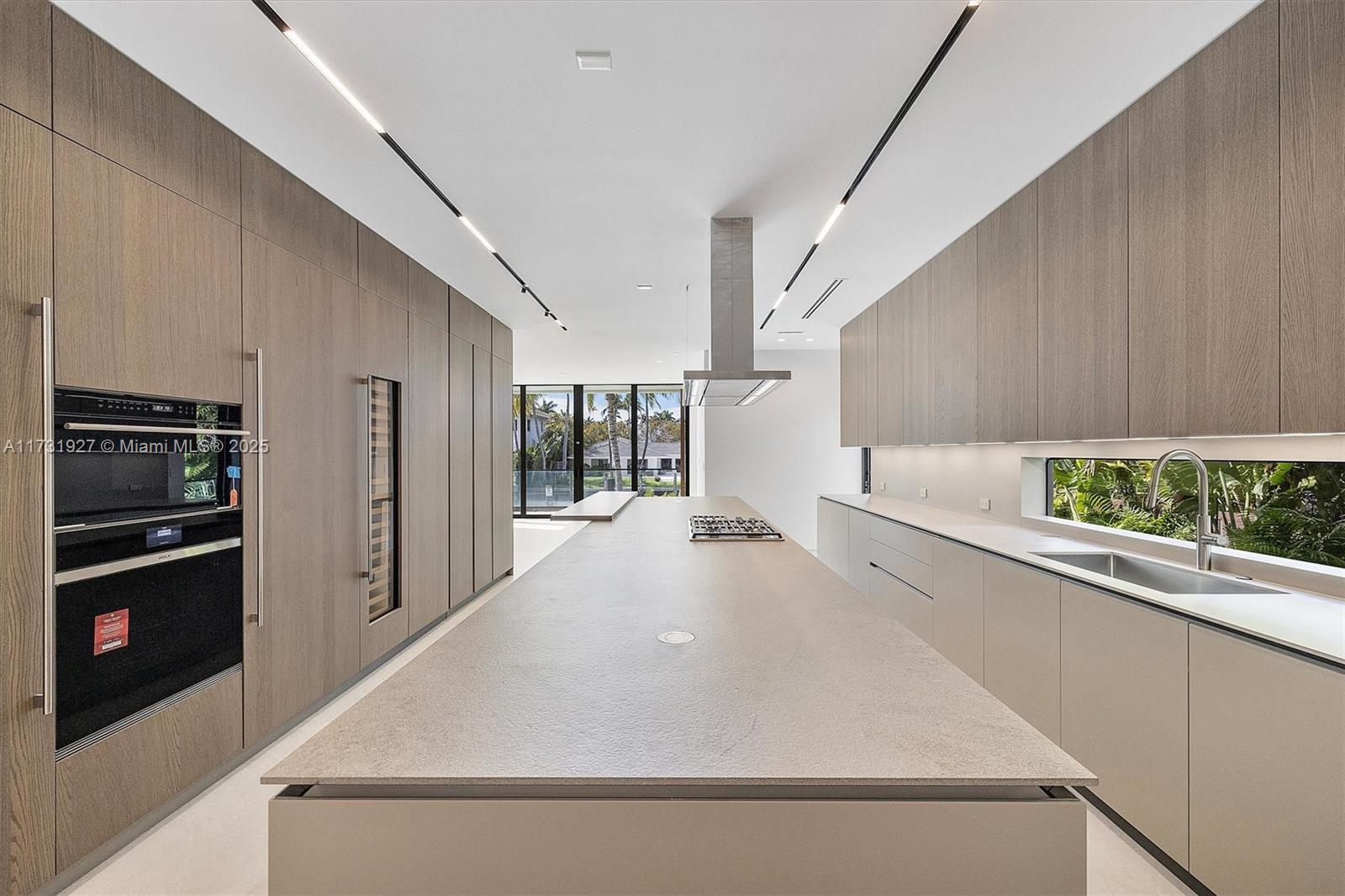 A modern kitchen features a long central island, wood-toned cabinetry, and a horizontal window backsplash — 960 Mariner Dr, Key Biscayne FL 33149, KEY BISCAYNE, FL