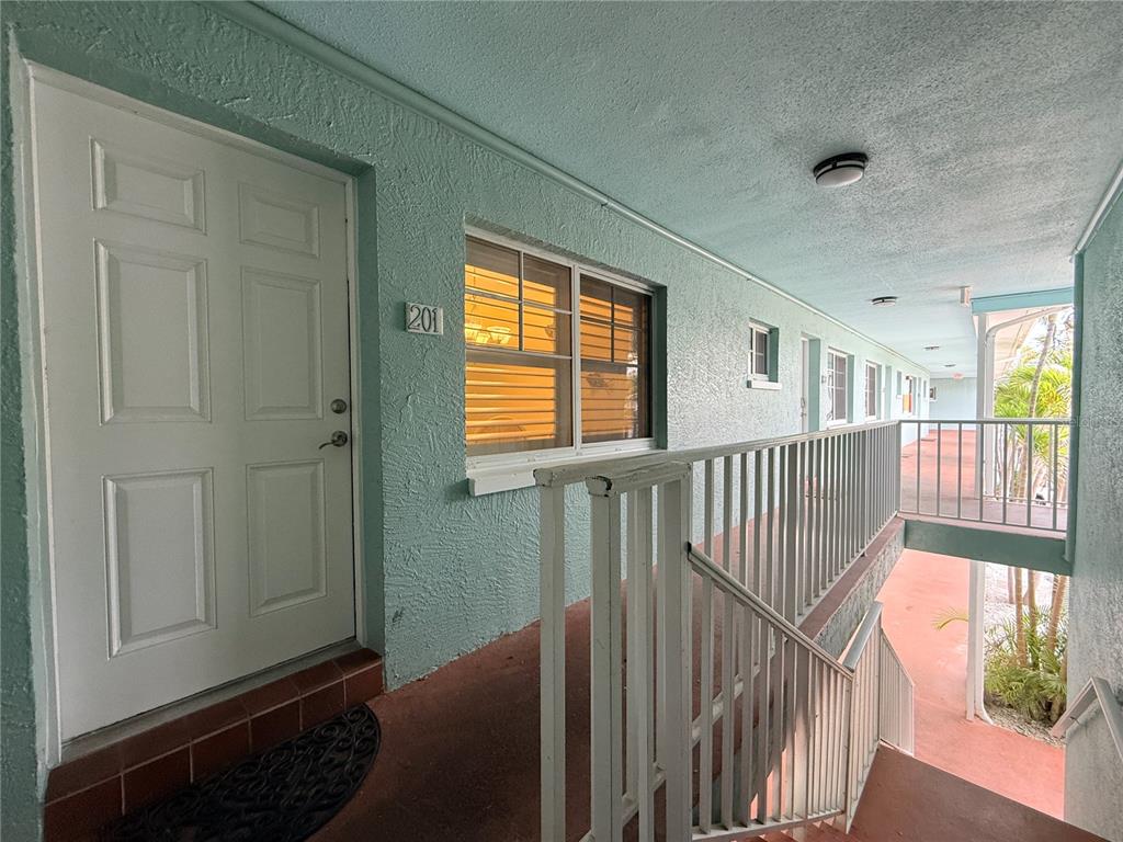 An exterior view of an apartment building walkway featuring a white door labeled 201 — 19417 GULF BOULEVARD UNIT D201, INDIAN ROCKS BEACH, FL