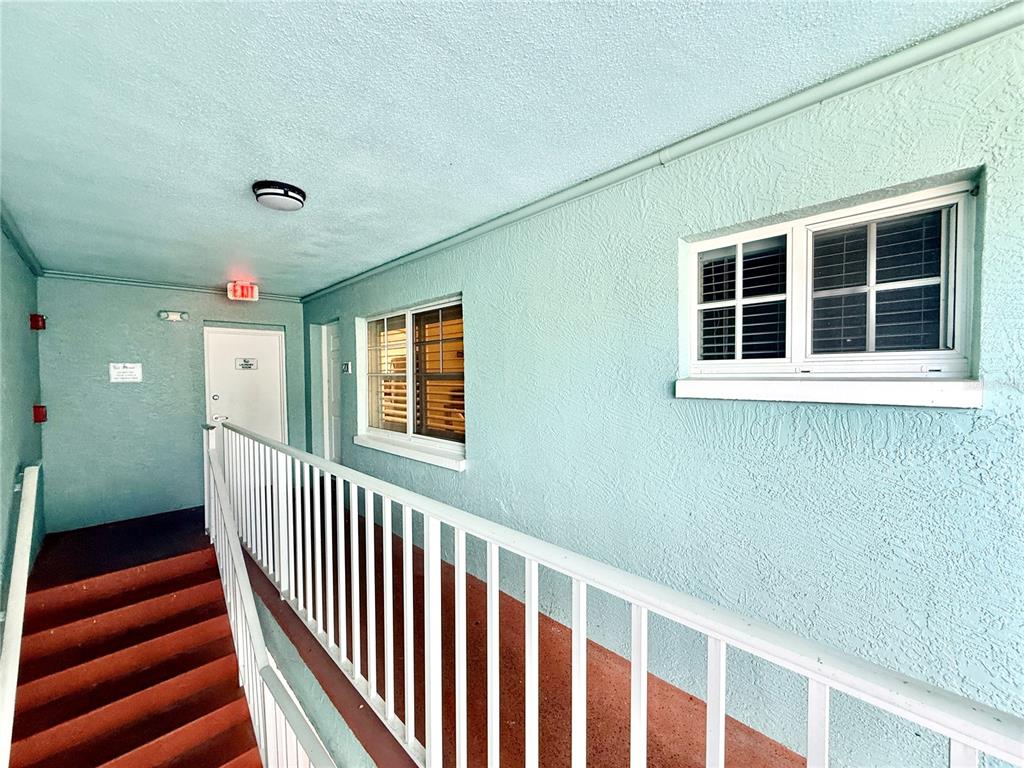 An outdoor apartment walkway features light blue stucco walls, a white railing, and red stairs — 19417 GULF BOULEVARD UNIT D201, INDIAN ROCKS BEACH, FL